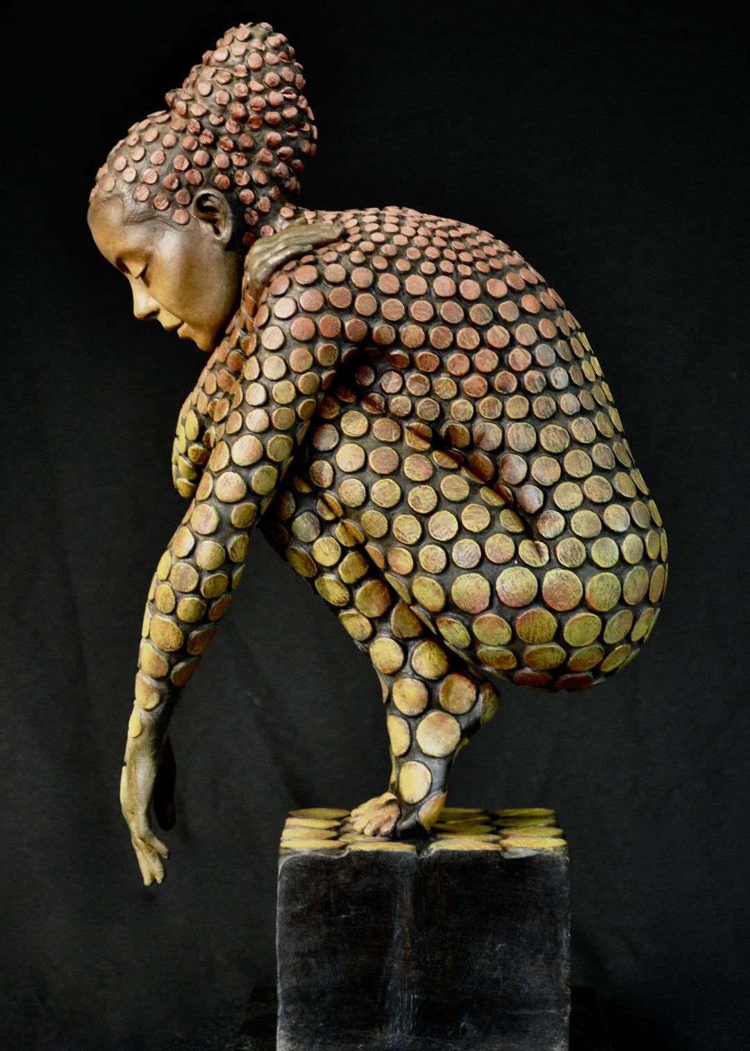 Bronze sculpture of a crouching female figure, her body covered in a gradual pattern of circular, sunset-toned discs that shift from warm reds to soft golds. She balances on a textured block, head bowed gently in a contemplative pose, with a dark backdrop emphasising the warmth and depth of the patina.