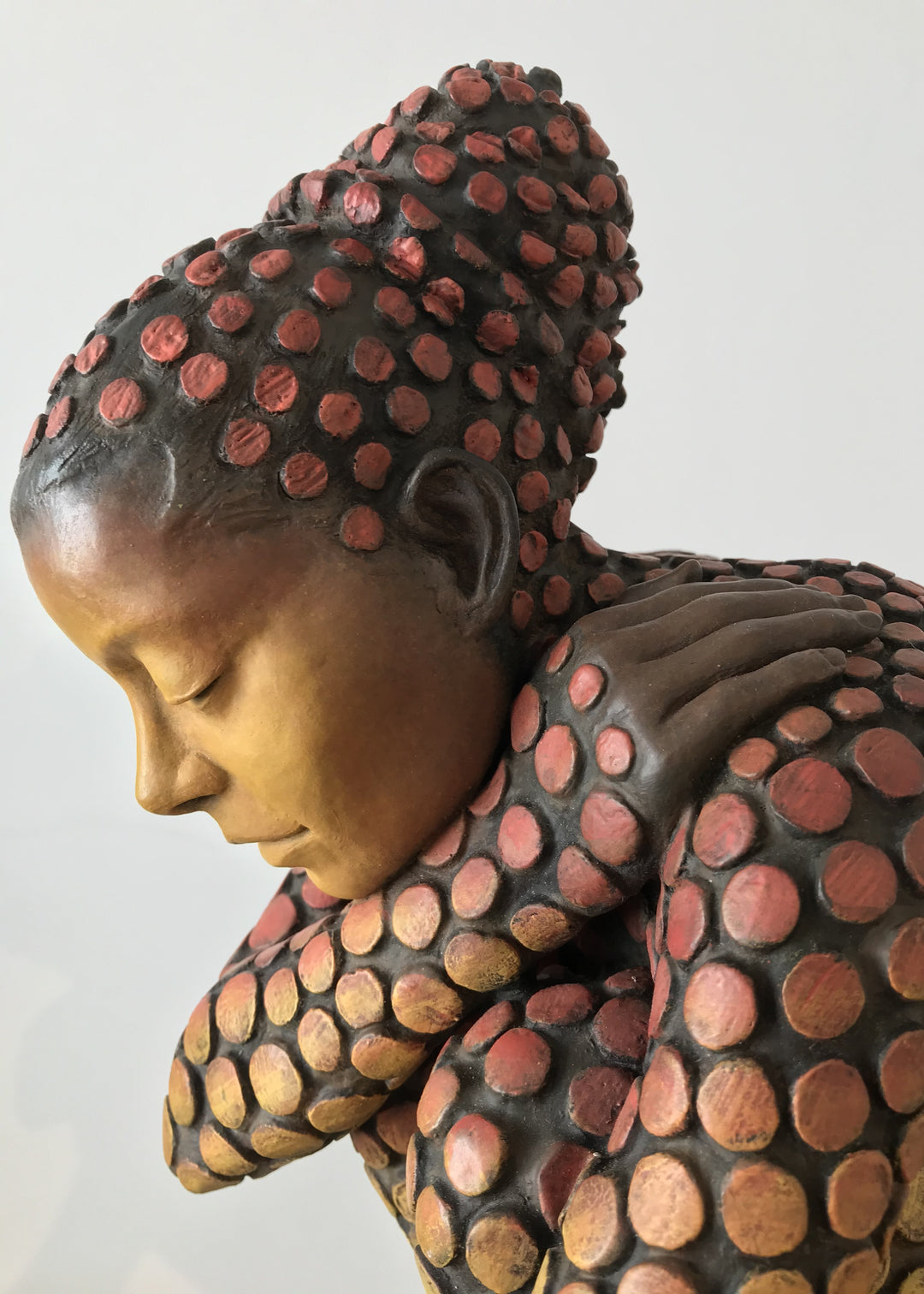 Bronze sculpture of a crouching female figure, her body covered in a gradual pattern of circular, sunset-toned discs that shift from warm reds to soft golds. She balances on a textured block, head bowed gently in a contemplative pose, with a dark backdrop emphasising the warmth and depth of the patina.