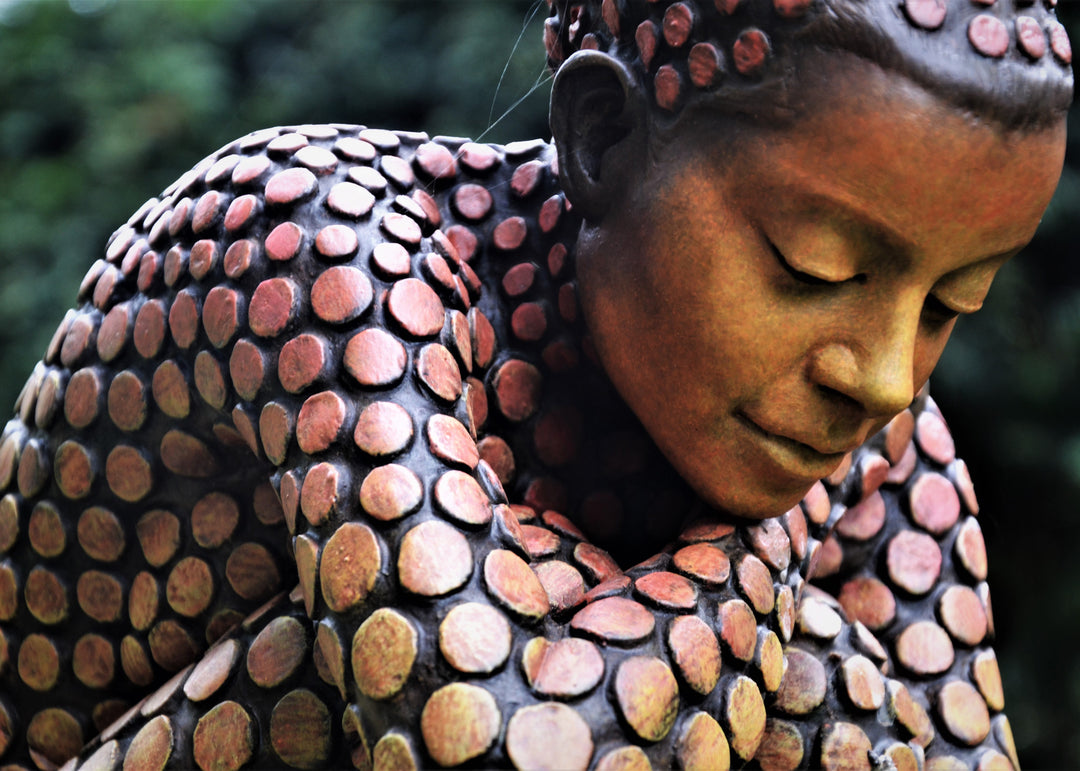 Bronze sculpture of a crouching female figure, her body covered in a gradual pattern of circular, sunset-toned discs that shift from warm reds to soft golds. She balances on a textured block, head bowed gently in a contemplative pose, with a dark backdrop emphasising the warmth and depth of the patina.