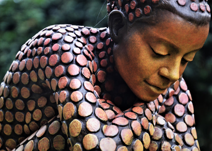 Bronze sculpture of a crouching female figure, her body covered in a gradual pattern of circular, sunset-toned discs that shift from warm reds to soft golds. She balances on a textured block, head bowed gently in a contemplative pose, with a dark backdrop emphasising the warmth and depth of the patina.