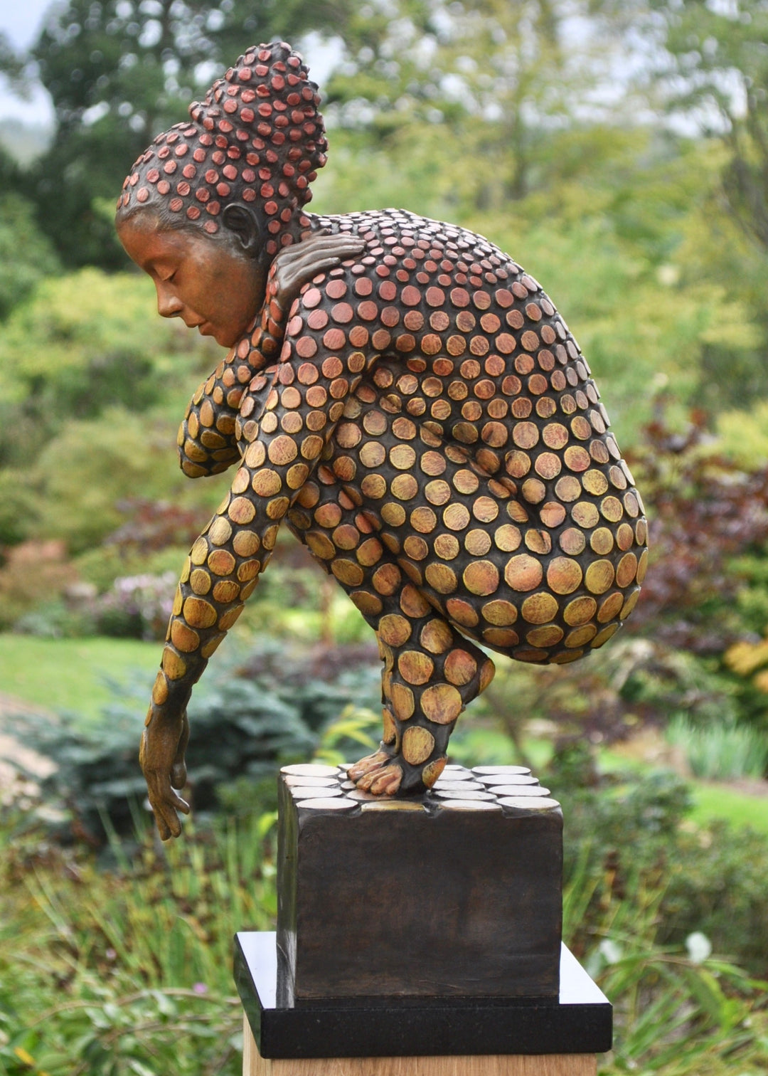 Bronze sculpture of a crouching female figure, her body covered in a gradual pattern of circular, sunset-toned discs that shift from warm reds to soft golds. She balances on a textured block, head bowed gently in a contemplative pose, with a dark backdrop emphasising the warmth and depth of the patina.
