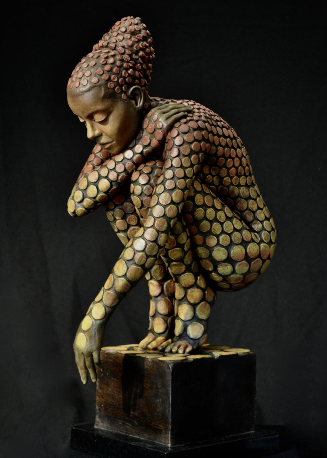 Bronze sculpture of a crouching female figure, her body covered in a gradual pattern of circular, sunset-toned discs that shift from warm reds to soft golds. She balances on a textured block, head bowed gently in a contemplative pose, with a dark backdrop emphasising the warmth and depth of the patina.