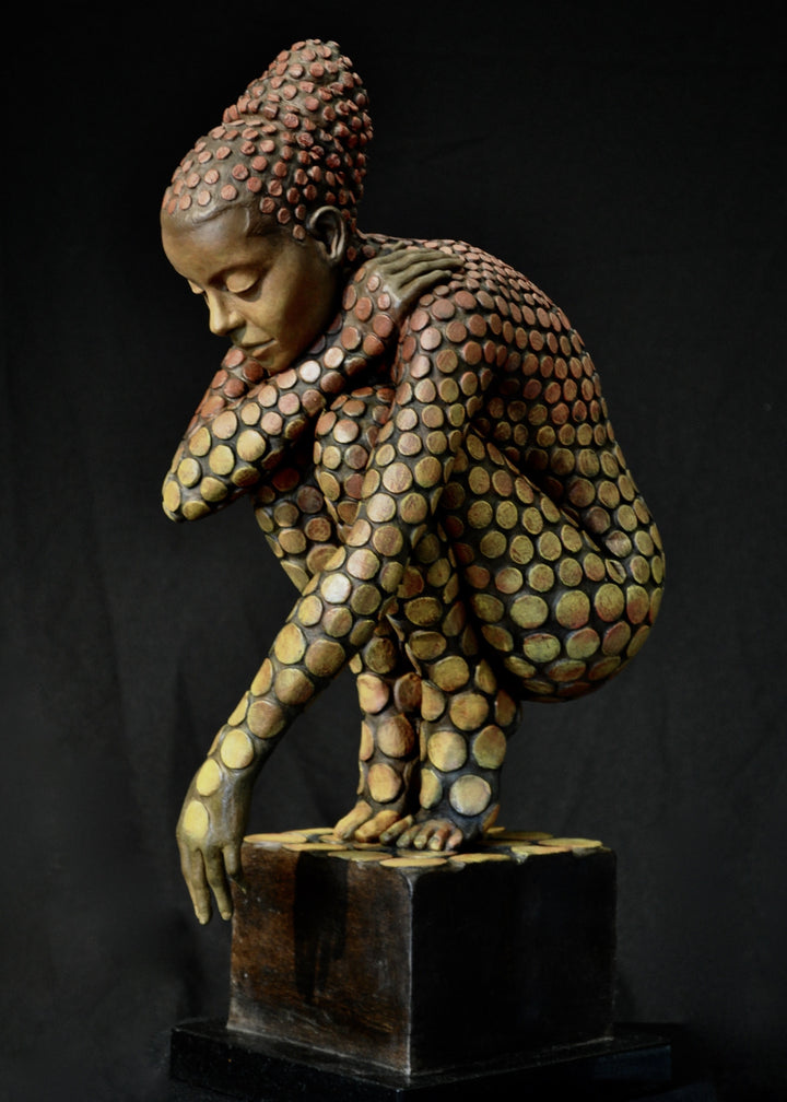 Bronze sculpture of a crouching female figure, her body covered in a gradual pattern of circular, sunset-toned discs that shift from warm reds to soft golds. She balances on a textured block, head bowed gently in a contemplative pose, with a dark backdrop emphasising the warmth and depth of the patina.