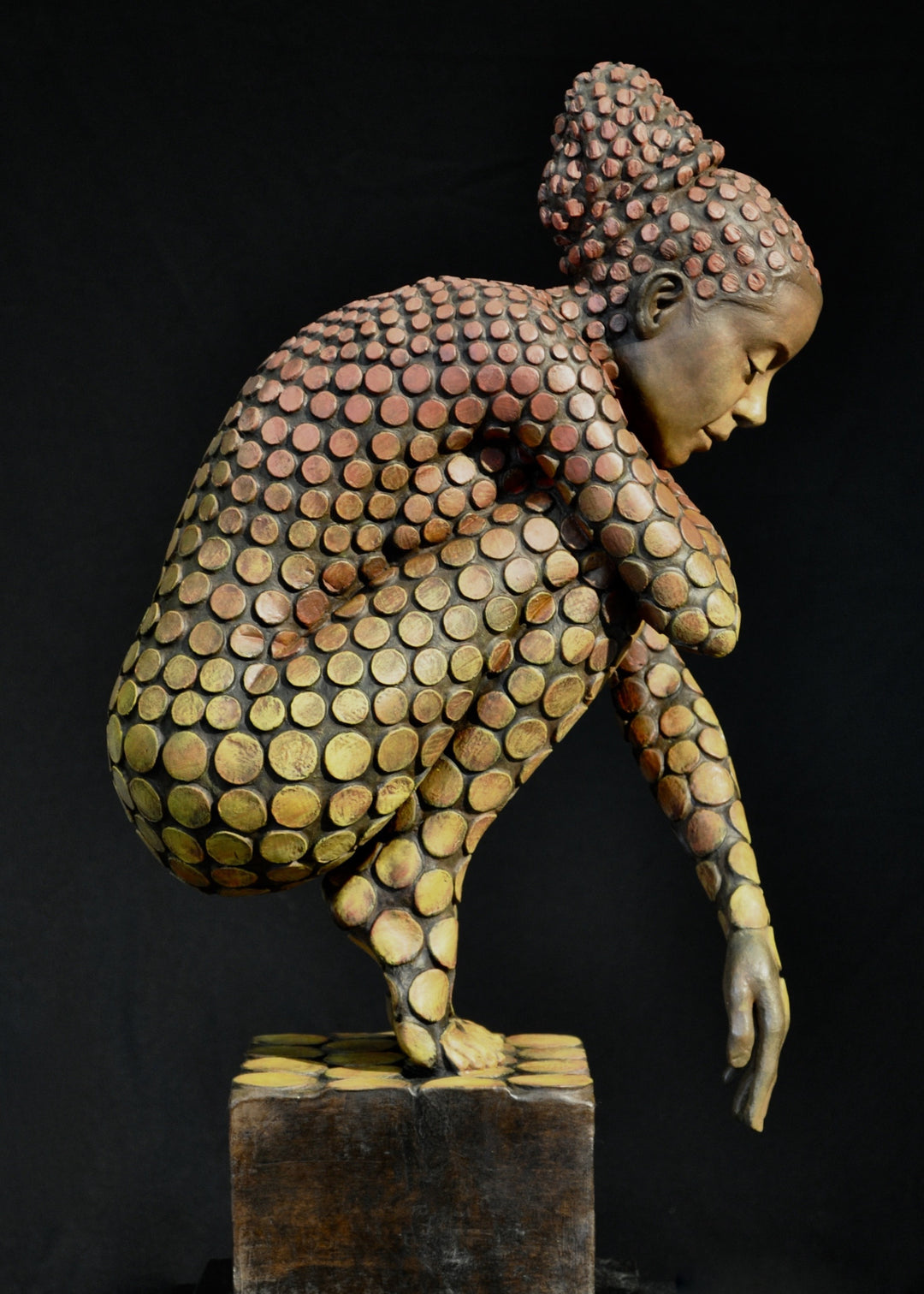 Bronze sculpture of a crouching female figure, her body covered in a gradual pattern of circular, sunset-toned discs that shift from warm reds to soft golds. She balances on a textured block, head bowed gently in a contemplative pose, with a dark backdrop emphasising the warmth and depth of the patina.