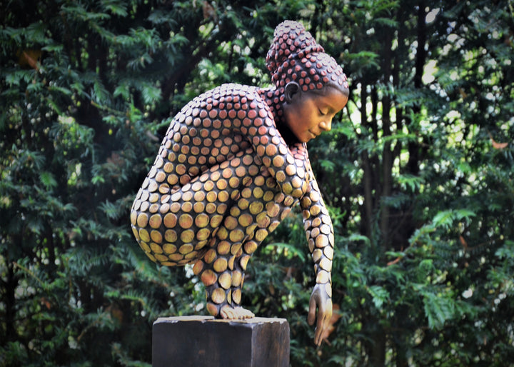 Bronze sculpture of a crouching female figure, her body covered in a gradual pattern of circular, sunset-toned discs that shift from warm reds to soft golds. She balances on a textured block, head bowed gently in a contemplative pose, with a dark backdrop emphasising the warmth and depth of the patina.