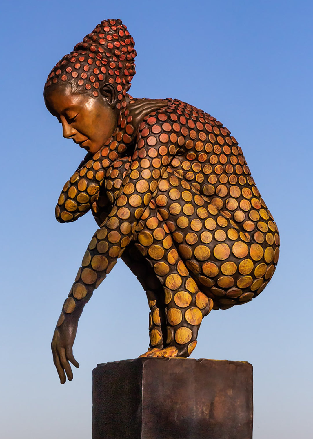 Bronze sculpture of a crouching female figure, her body covered in a gradual pattern of circular, sunset-toned discs that shift from warm reds to soft golds. She balances on a textured block, head bowed gently in a contemplative pose, with a dark backdrop emphasising the warmth and depth of the patina.