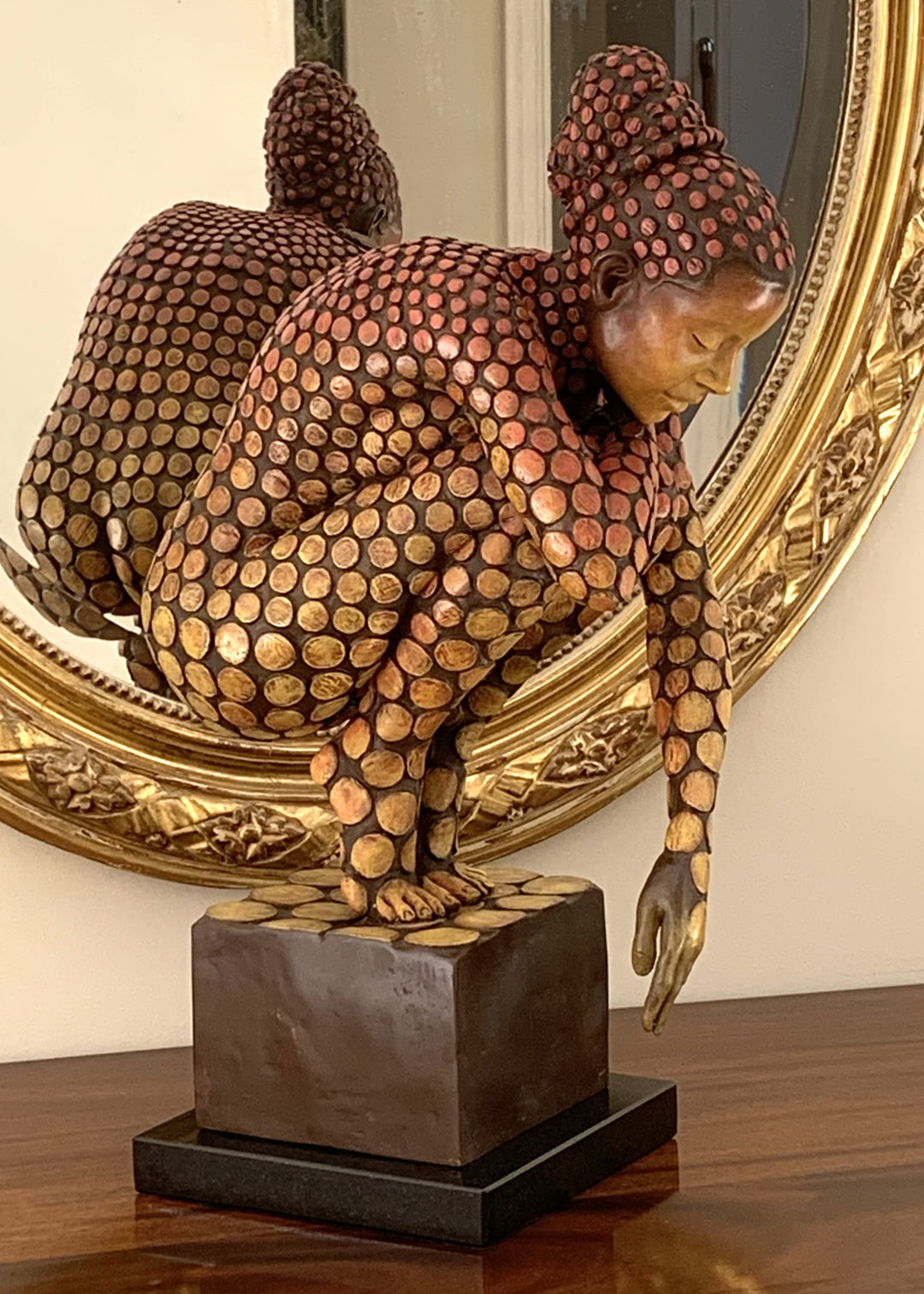 Bronze sculpture of a crouching female figure, her body covered in a gradual pattern of circular, sunset-toned discs that shift from warm reds to soft golds. She balances on a textured block, head bowed gently in a contemplative pose, with a dark backdrop emphasising the warmth and depth of the patina.