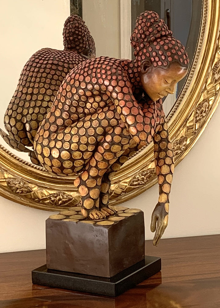 Bronze sculpture of a crouching female figure, her body covered in a gradual pattern of circular, sunset-toned discs that shift from warm reds to soft golds. She balances on a textured block, head bowed gently in a contemplative pose, with a dark backdrop emphasising the warmth and depth of the patina.