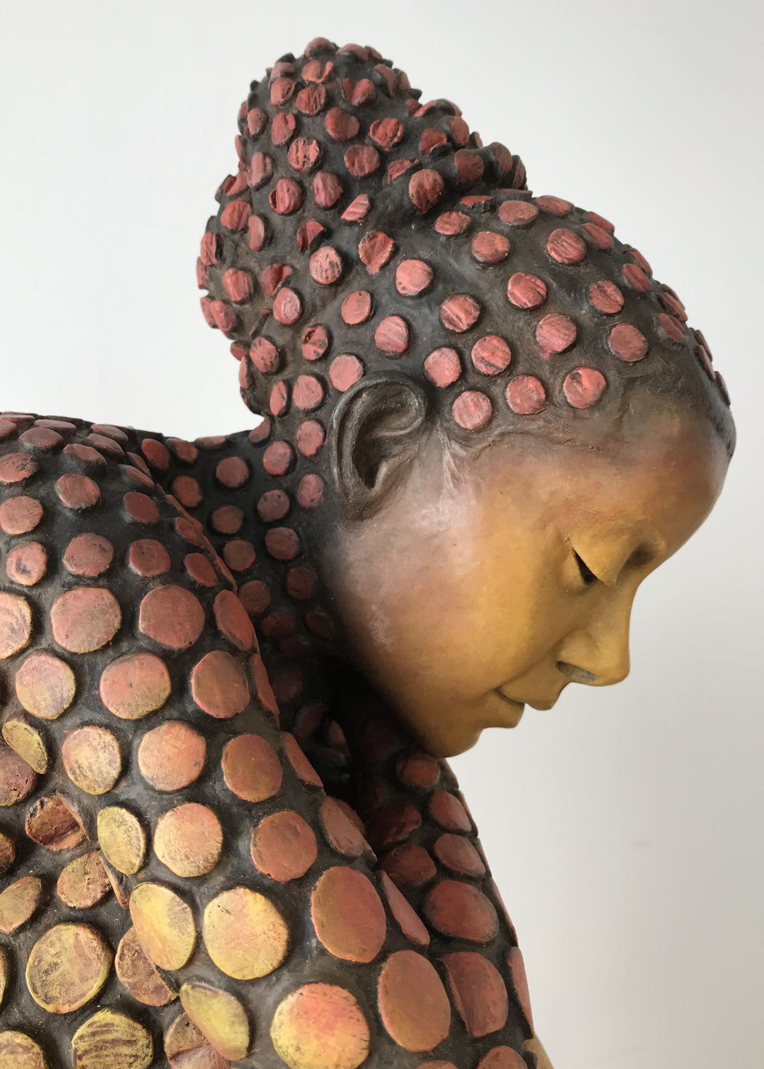 Bronze sculpture of a crouching female figure, her body covered in a gradual pattern of circular, sunset-toned discs that shift from warm reds to soft golds. She balances on a textured block, head bowed gently in a contemplative pose, with a dark backdrop emphasising the warmth and depth of the patina.