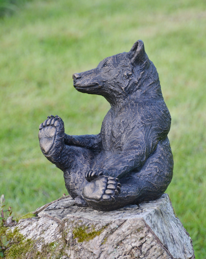 Playful bear cub sculpture sitting upright on a tree stump, with textured dark surface detail and a friendly expression, set against a blurred garden background.