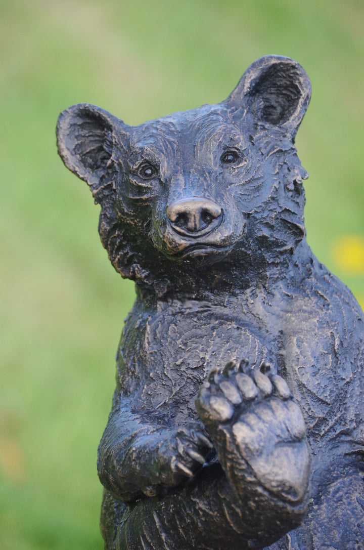 Playful bear cub sculpture sitting upright on a tree stump, with textured dark surface detail and a friendly expression, set against a blurred garden background.