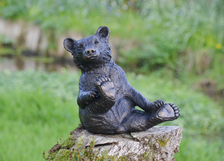 Playful bear cub sculpture sitting upright on a tree stump, with textured dark surface detail and a friendly expression, set against a blurred garden background.