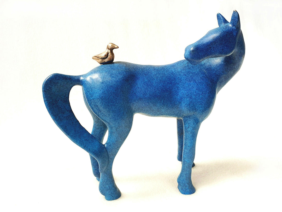 Sculptural artwork of a stylised blue horse with smooth curves and minimalist form, featuring a small bronze bird perched on its back. The ceramic surface has a mottled cobalt-blue glaze, creating a rich, vibrant finish against a white backdrop.