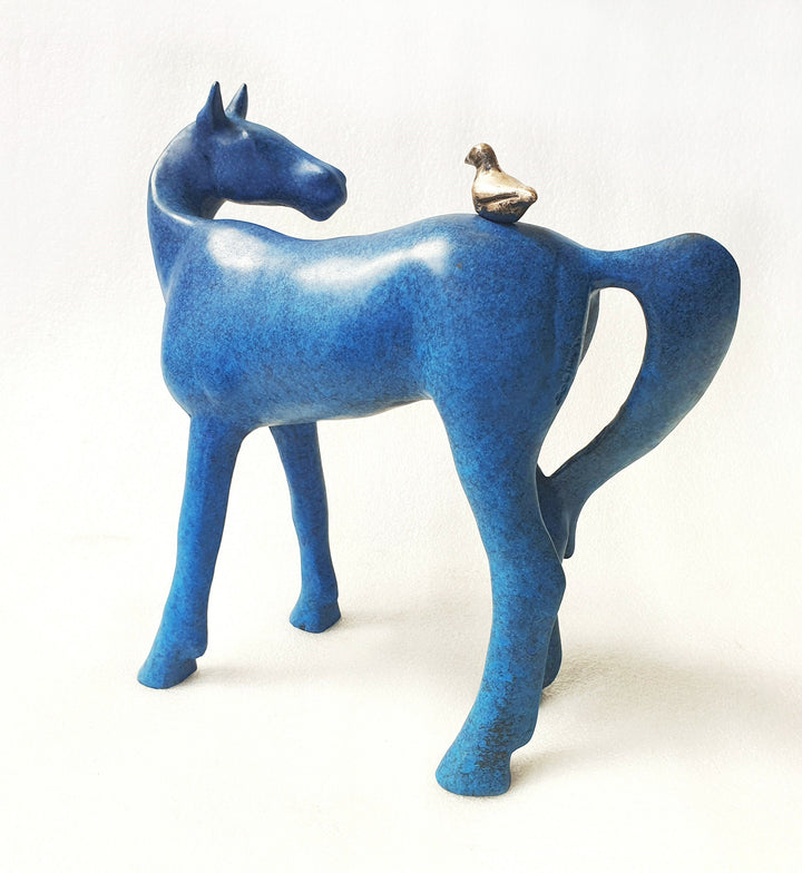 Sculptural artwork of a stylised blue horse with smooth curves and minimalist form, featuring a small bronze bird perched on its back. The ceramic surface has a mottled cobalt-blue glaze, creating a rich, vibrant finish against a white backdrop.
