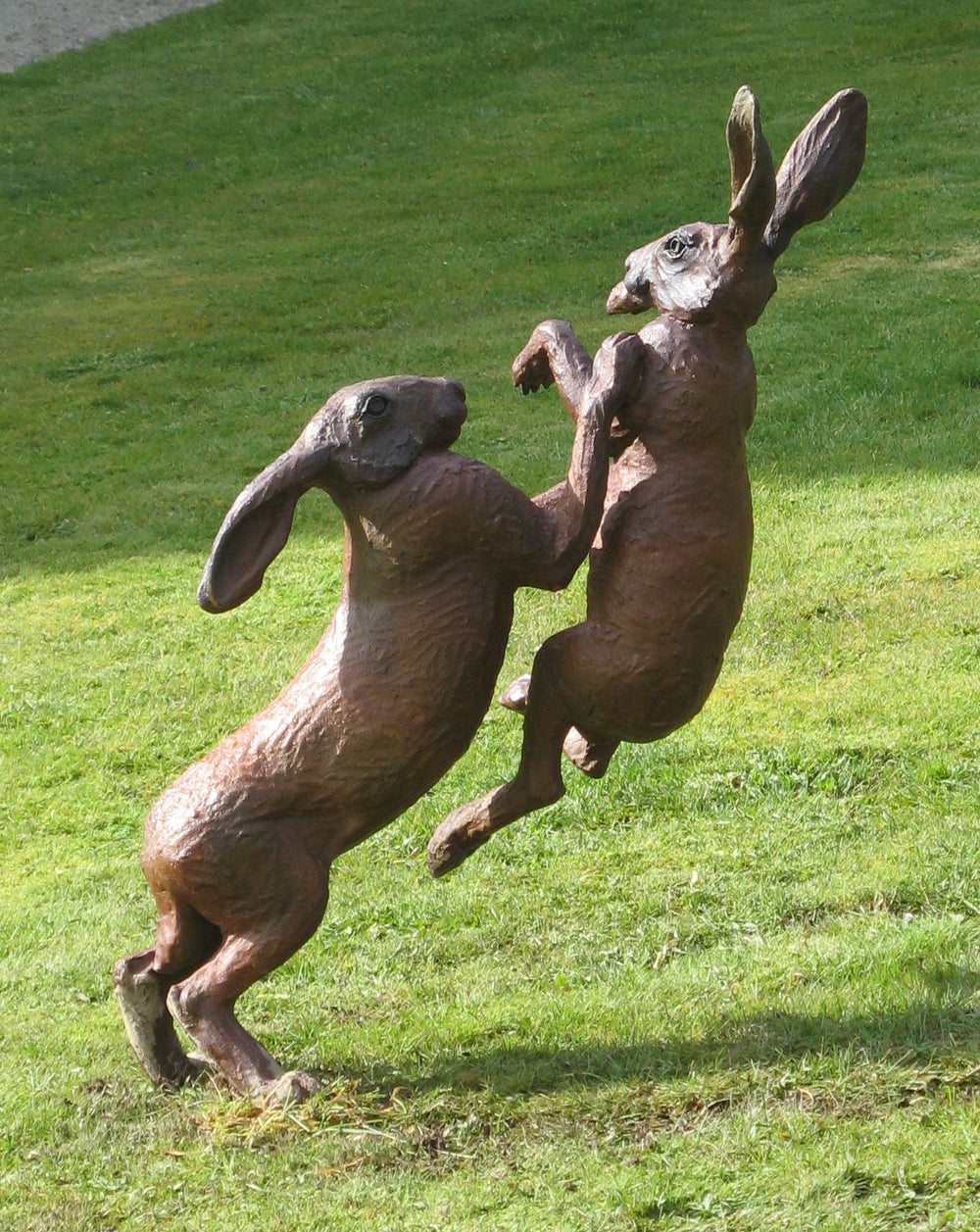 Bronze sculpture of two hares mid-boxing, captured in an energetic and dynamic pose on a grassy lawn. The detailing emphasises the muscular forms and textured fur, with one hare slightly airborne as they engage in playful combat.