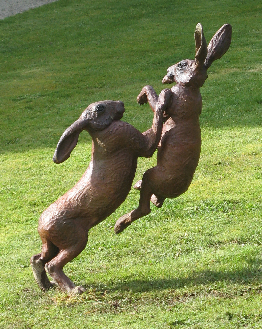 Bronze sculpture of two hares mid-boxing, captured in an energetic and dynamic pose on a grassy lawn. The detailing emphasises the muscular forms and textured fur, with one hare slightly airborne as they engage in playful combat.