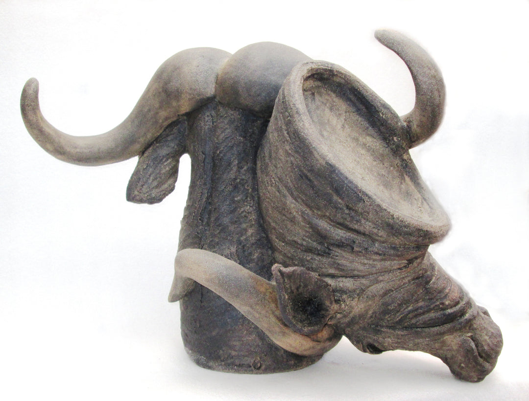 Sculptural ceramic of two buffalo heads intertwined, crafted in textured stoneware with naturalistic detail and expressive curvature. One head faces forward while the other tilts downward, their horns looping in dynamic, sculptural tension against a neutral background.