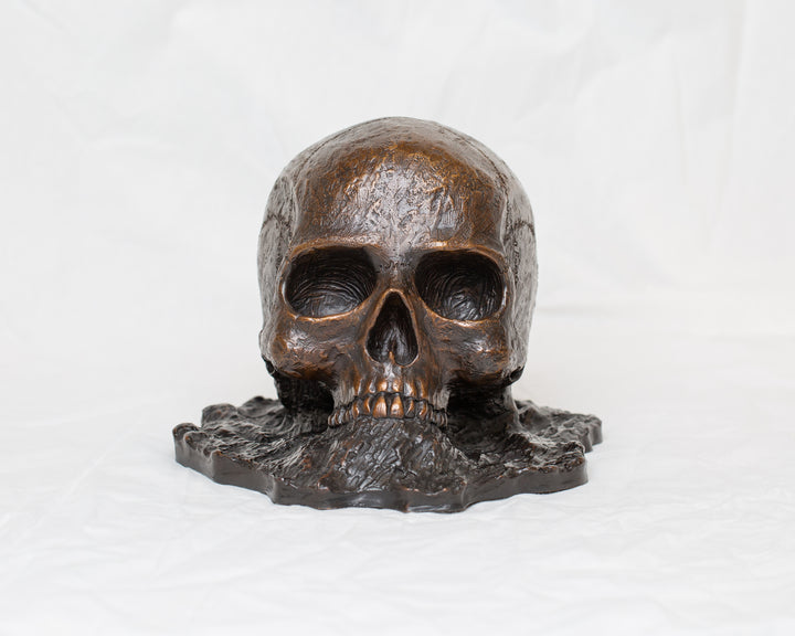 Remember Death bronze skull sculpture by Tom Saxton emerging from textured bronze ground, detailed human skull memento mori sculpture in rich dark bronze, warm copper-brown tones and deep shadowed cavities, contemporary philosophical bronze artwork