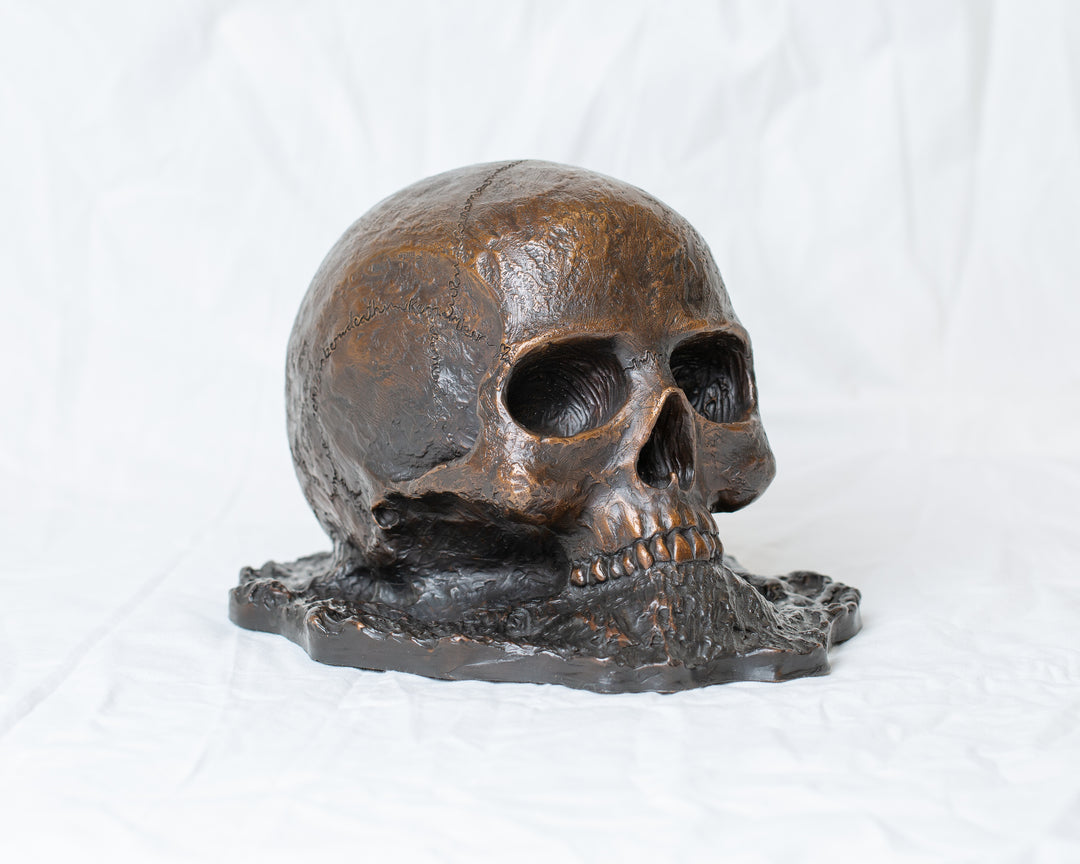 Remember Death bronze skull sculpture by Tom Saxton emerging from textured bronze ground, detailed human skull memento mori sculpture in rich dark bronze, warm copper-brown tones and deep shadowed cavities, contemporary philosophical bronze artwork