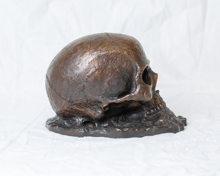 Remember Death bronze skull sculpture by Tom Saxton emerging from textured bronze ground, detailed human skull memento mori sculpture in rich dark bronze, warm copper-brown tones and deep shadowed cavities, contemporary philosophical bronze artwork