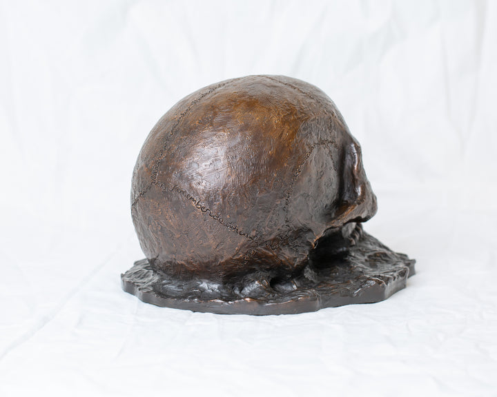 Remember Death bronze skull sculpture by Tom Saxton emerging from textured bronze ground, detailed human skull memento mori sculpture in rich dark bronze, warm copper-brown tones and deep shadowed cavities, contemporary philosophical bronze artwork