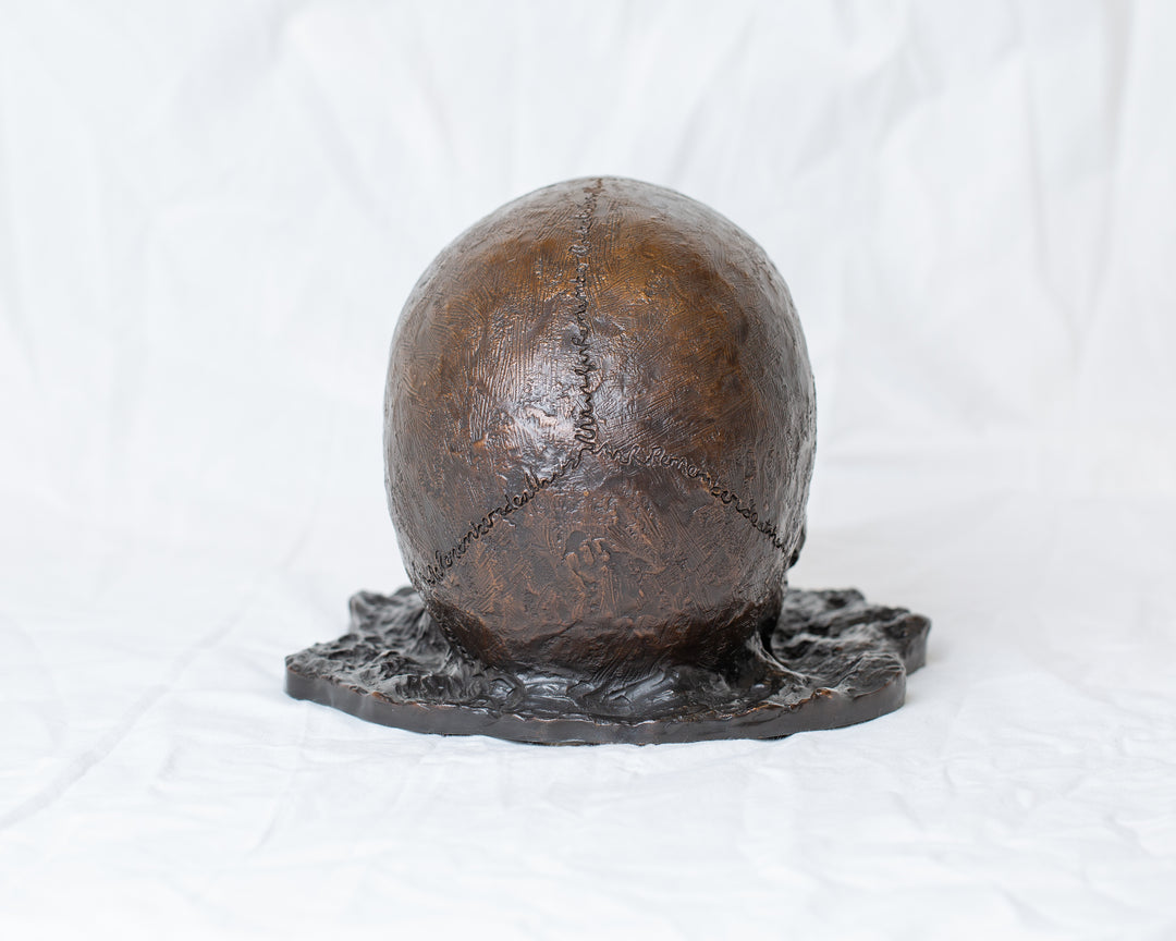 Remember Death bronze skull sculpture by Tom Saxton emerging from textured bronze ground, detailed human skull memento mori sculpture in rich dark bronze, warm copper-brown tones and deep shadowed cavities, contemporary philosophical bronze artwork