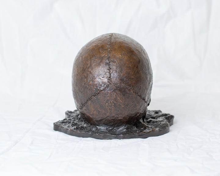 Remember Death bronze skull sculpture by Tom Saxton emerging from textured bronze ground, detailed human skull memento mori sculpture in rich dark bronze, warm copper-brown tones and deep shadowed cavities, contemporary philosophical bronze artwork