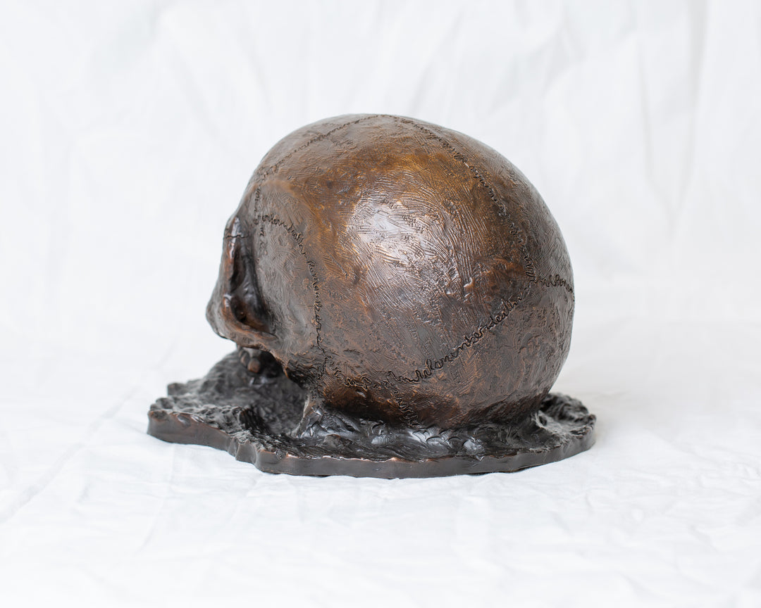 Remember Death bronze skull sculpture by Tom Saxton emerging from textured bronze ground, detailed human skull memento mori sculpture in rich dark bronze, warm copper-brown tones and deep shadowed cavities, contemporary philosophical bronze artwork