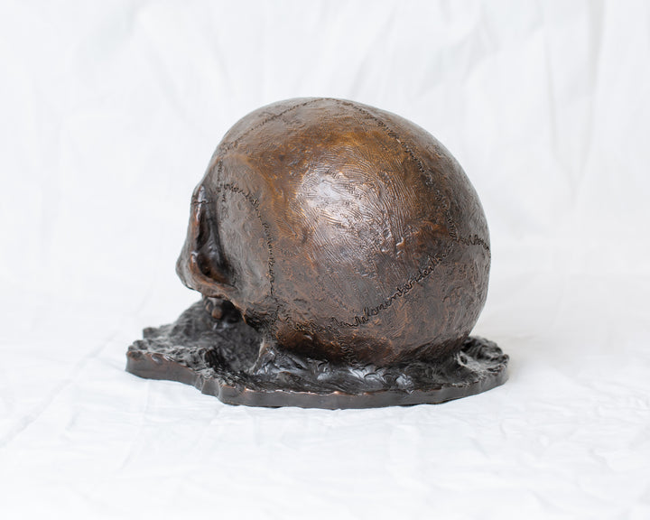 Remember Death bronze skull sculpture by Tom Saxton emerging from textured bronze ground, detailed human skull memento mori sculpture in rich dark bronze, warm copper-brown tones and deep shadowed cavities, contemporary philosophical bronze artwork