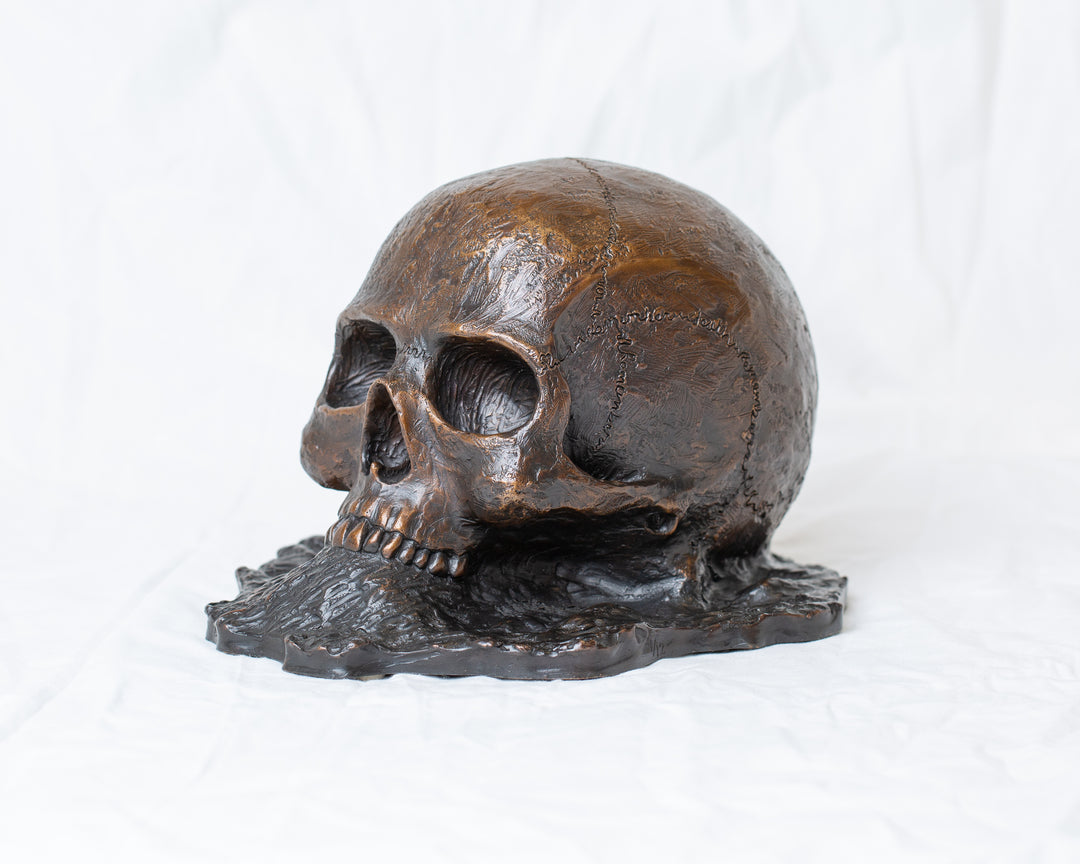 Remember Death bronze skull sculpture by Tom Saxton emerging from textured bronze ground, detailed human skull memento mori sculpture in rich dark bronze, warm copper-brown tones and deep shadowed cavities, contemporary philosophical bronze artwork