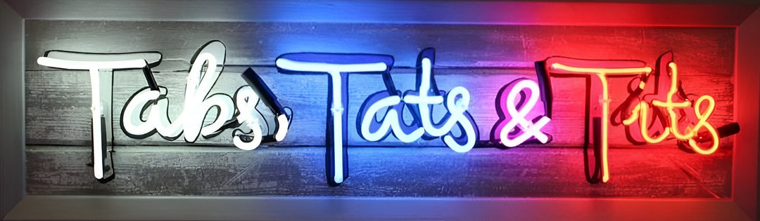 Neon artwork by Matthew Bracey spelling out the phrase "Tabs, Tats & Tits" in bold, playful script. The piece features vibrant white, blue, pink, and yellow neon against a rustic wooden background, with the final word illuminated in glowing red hues, creating a cheeky and irreverent statement piece.