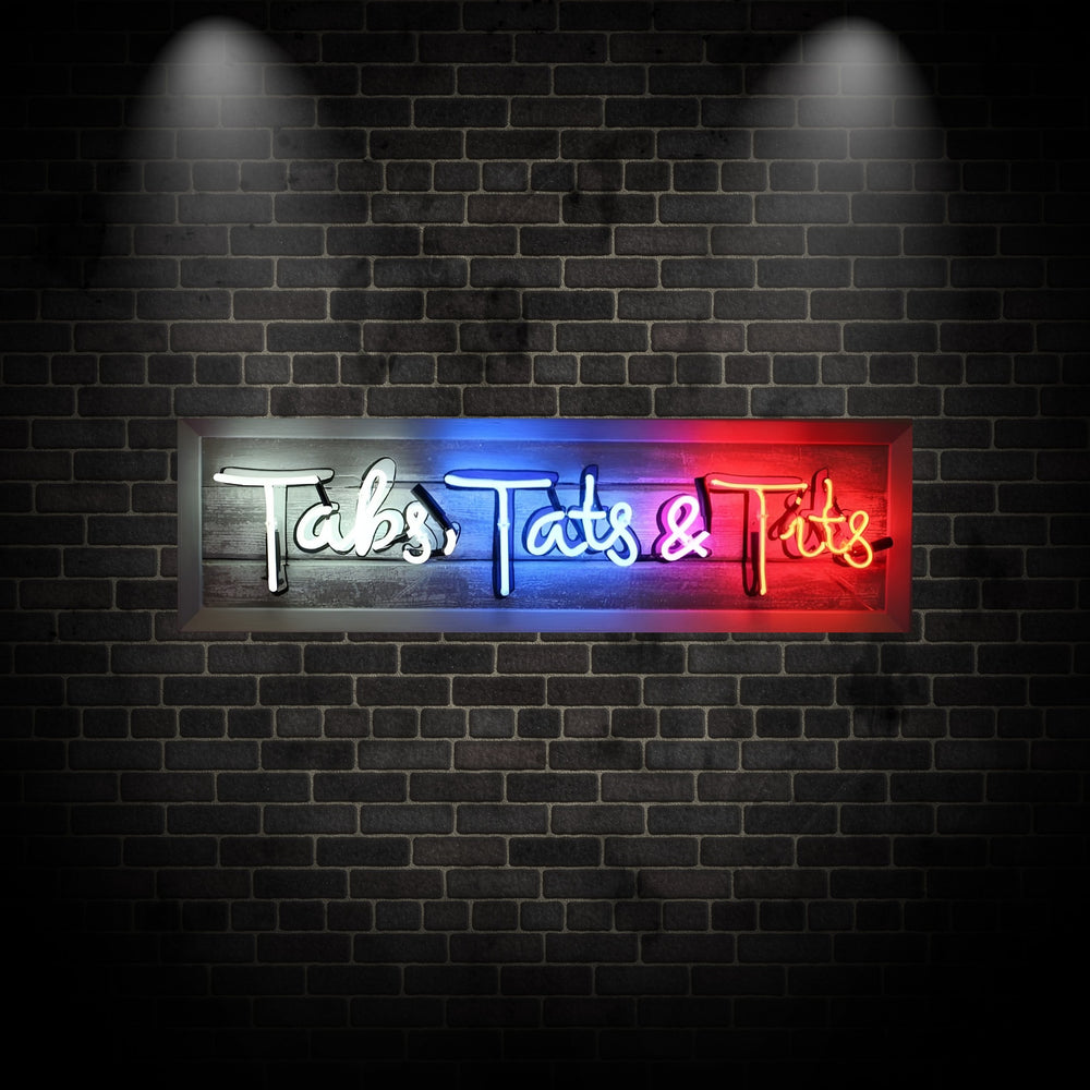 Neon artwork by Matthew Bracey spelling out the phrase "Tabs, Tats & Tits" in bold, playful script. The piece features vibrant white, blue, pink, and yellow neon against a rustic wooden background, with the final word illuminated in glowing red hues, creating a cheeky and irreverent statement piece.