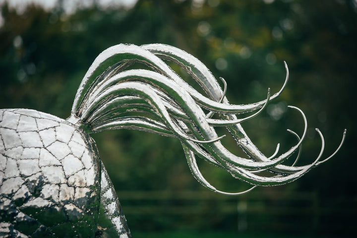 ‘Leaping Horse’ by Michael Turner, a unique, one-of-one stainless steel equestrian sculpture depicting a magnificent life-size horse leaping forward with its front legs raised and hind legs extended. Handcrafted from marine-grade stainless steel, the sculpture features intricately welded metallic plates, a powerful muscular body, flowing mane and sweeping tail. Its polished silver finish reflects the surrounding landscape, while the dynamic pose captures the strength, elegance and movement of a horse in ful