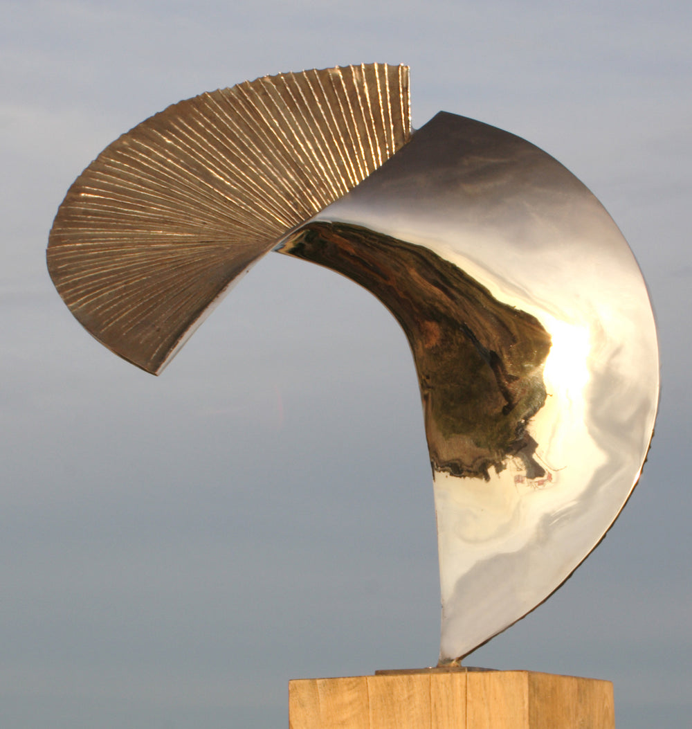 Polished bronze sculpture titled Talisman by Thomas Joynes, featuring a fan-like composition with two asymmetrical, opposing arcs. One half is mirror-polished to a high gleam, reflecting the surrounding environment, while the other is deeply textured with radiating linear grooves and a rich dark patina. Mounted on a wooden plinth, the piece elegantly balances tension and flow in a compact, tabletop scale.