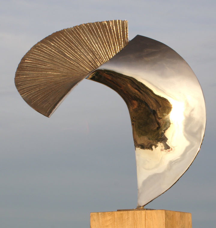 Polished bronze sculpture titled Talisman by Thomas Joynes, featuring a fan-like composition with two asymmetrical, opposing arcs. One half is mirror-polished to a high gleam, reflecting the surrounding environment, while the other is deeply textured with radiating linear grooves and a rich dark patina. Mounted on a wooden plinth, the piece elegantly balances tension and flow in a compact, tabletop scale.