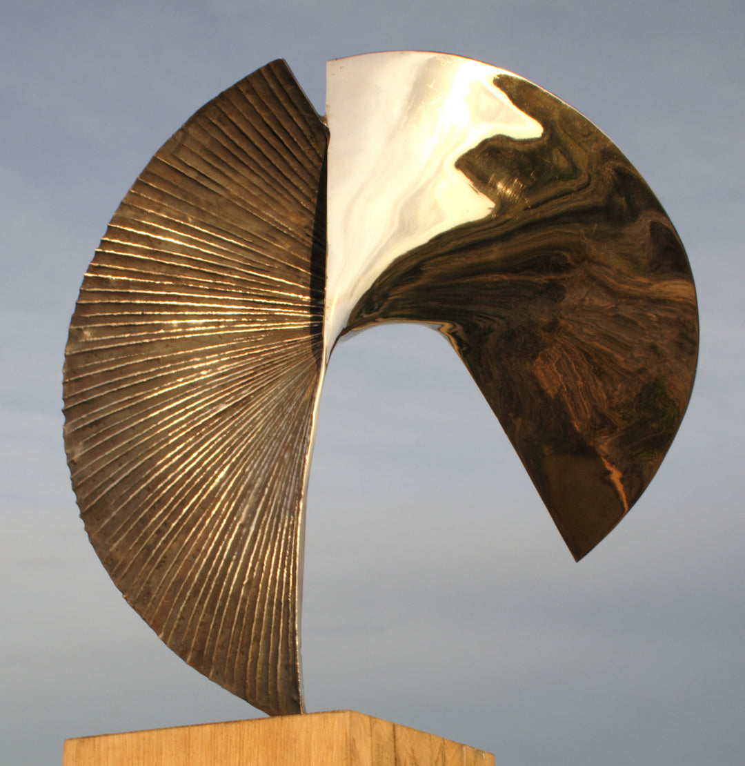 Polished bronze sculpture titled Talisman by Thomas Joynes, featuring a fan-like composition with two asymmetrical, opposing arcs. One half is mirror-polished to a high gleam, reflecting the surrounding environment, while the other is deeply textured with radiating linear grooves and a rich dark patina. Mounted on a wooden plinth, the piece elegantly balances tension and flow in a compact, tabletop scale.