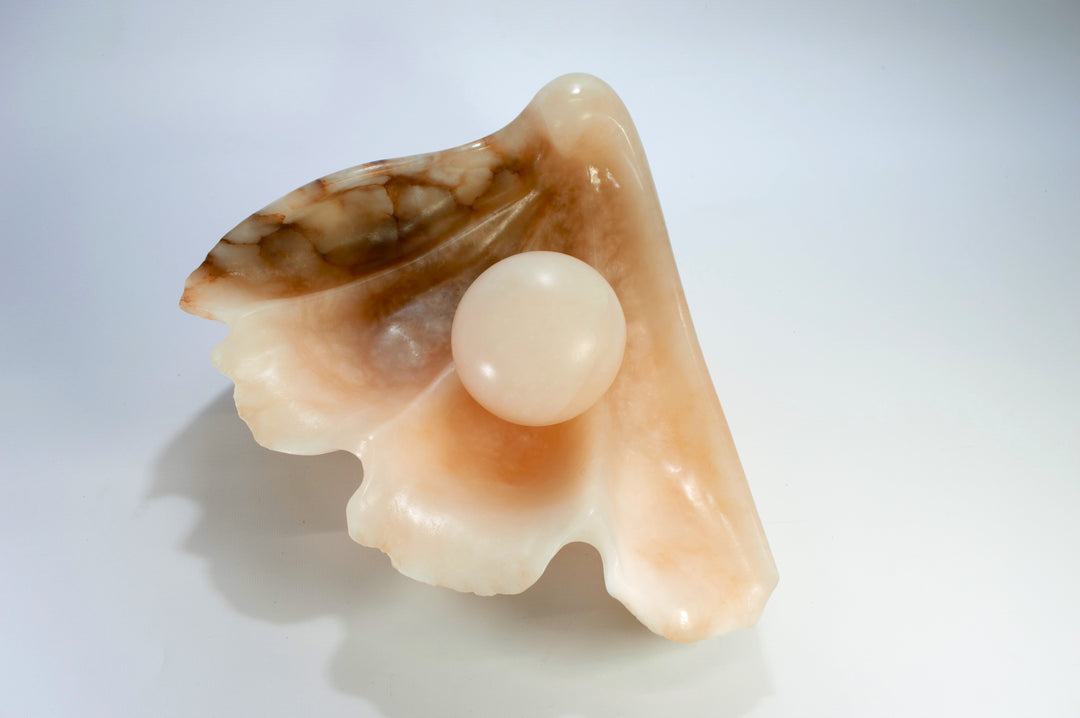 Deborah Harrison sculpture Tear of Joy, a one-off alabaster artwork resembling a shell-like vessel cradling a smooth alabaster sphere. The stone’s warm peach, cream, and brown veining creates a natural marbled effect, suggesting purity, renewal, and the symbolic form of a pearl within a shell.
