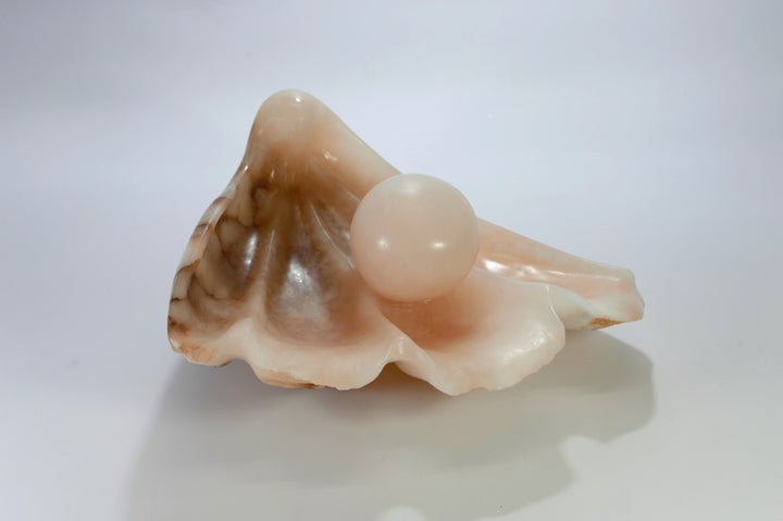 Deborah Harrison sculpture Tear of Joy, a one-off alabaster artwork resembling a shell-like vessel cradling a smooth alabaster sphere. The stone’s warm peach, cream, and brown veining creates a natural marbled effect, suggesting purity, renewal, and the symbolic form of a pearl within a shell.