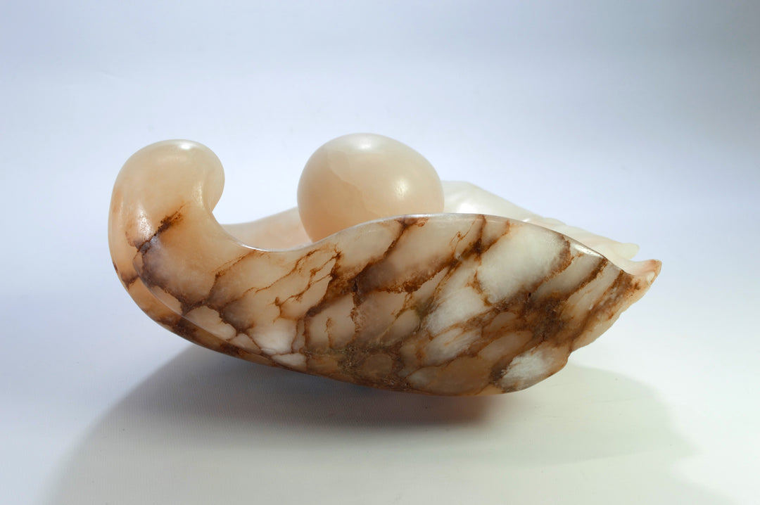 Deborah Harrison sculpture Tear of Joy, a one-off alabaster artwork resembling a shell-like vessel cradling a smooth alabaster sphere. The stone’s warm peach, cream, and brown veining creates a natural marbled effect, suggesting purity, renewal, and the symbolic form of a pearl within a shell.