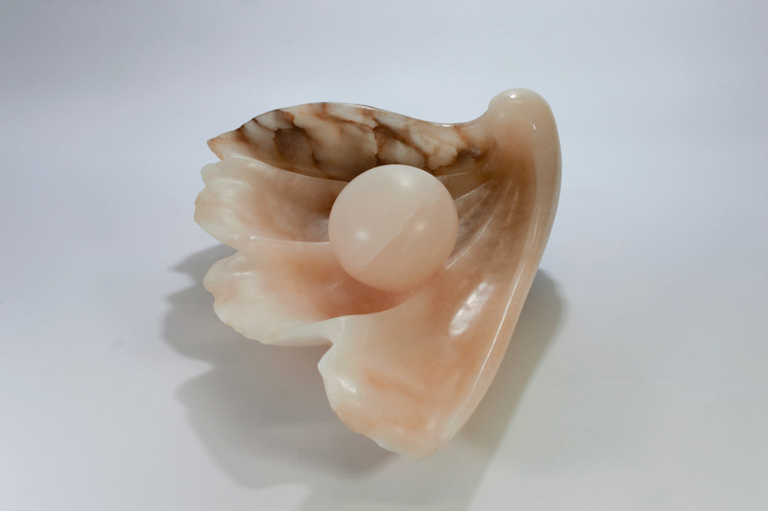 Deborah Harrison sculpture Tear of Joy, a one-off alabaster artwork resembling a shell-like vessel cradling a smooth alabaster sphere. The stone’s warm peach, cream, and brown veining creates a natural marbled effect, suggesting purity, renewal, and the symbolic form of a pearl within a shell.