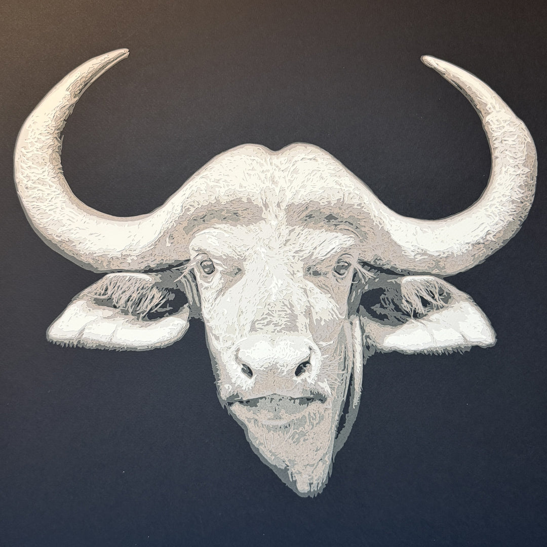 Hand-cut paper artwork of a Cape buffalo head with sweeping horns, crafted from eight layers in tones of ivory, stone grey, and warm taupe. The calm, commanding gaze captures strength and resilience, framed in black wood with UV-protective art glass.