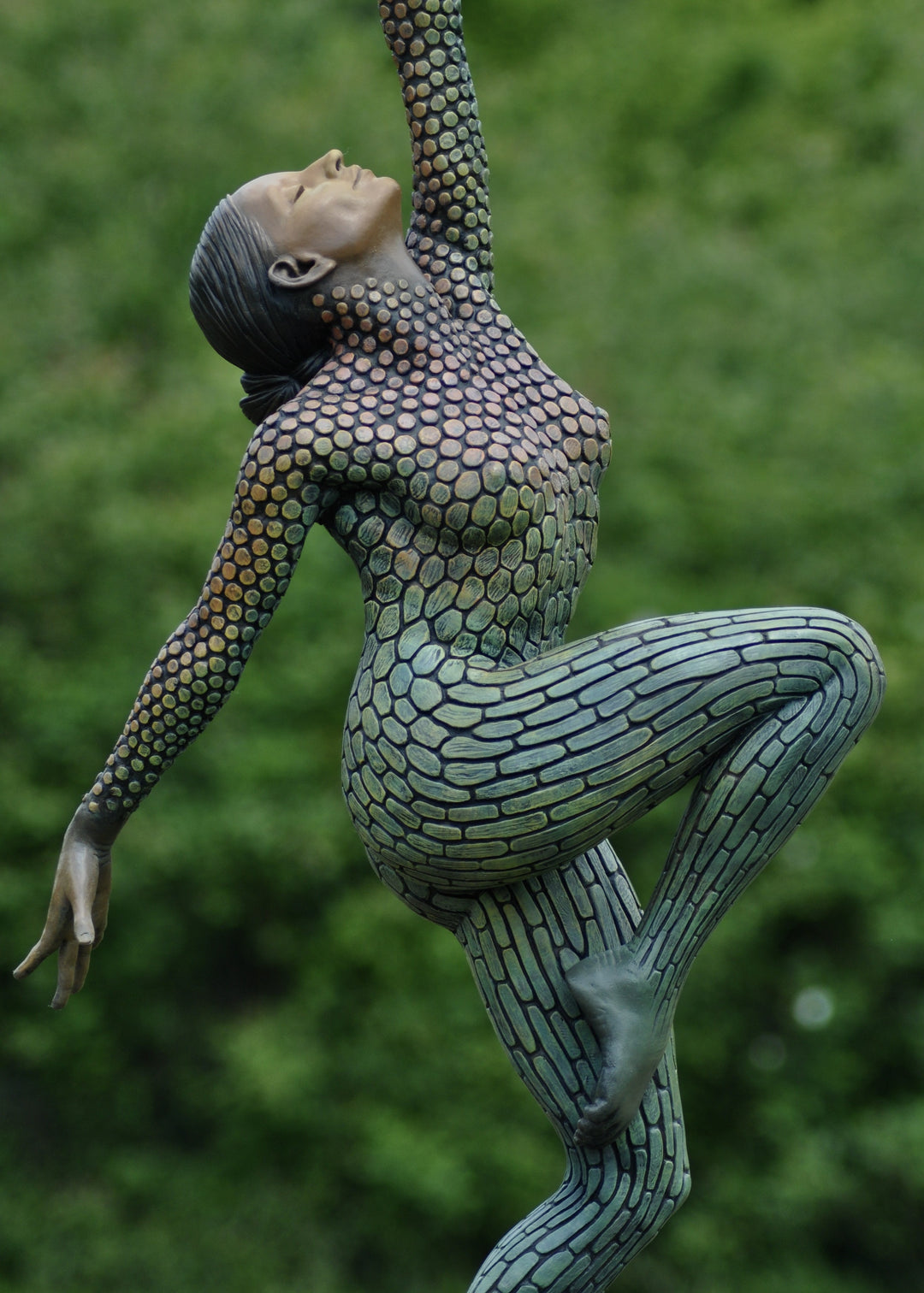 Life-size bronze sculpture of a woman balancing on one foot, her body covered in hand-painted mosaic-like textured scales that shift from earthy tones to green and blue patinas. She reaches upward with eyes closed, captured in a graceful, expressive pose against an outdoor garden backdrop.
