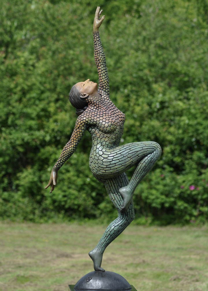 Life-size bronze sculpture of a woman balancing on one foot, her body covered in hand-painted mosaic-like textured scales that shift from earthy tones to green and blue patinas. She reaches upward with eyes closed, captured in a graceful, expressive pose against an outdoor garden backdrop.