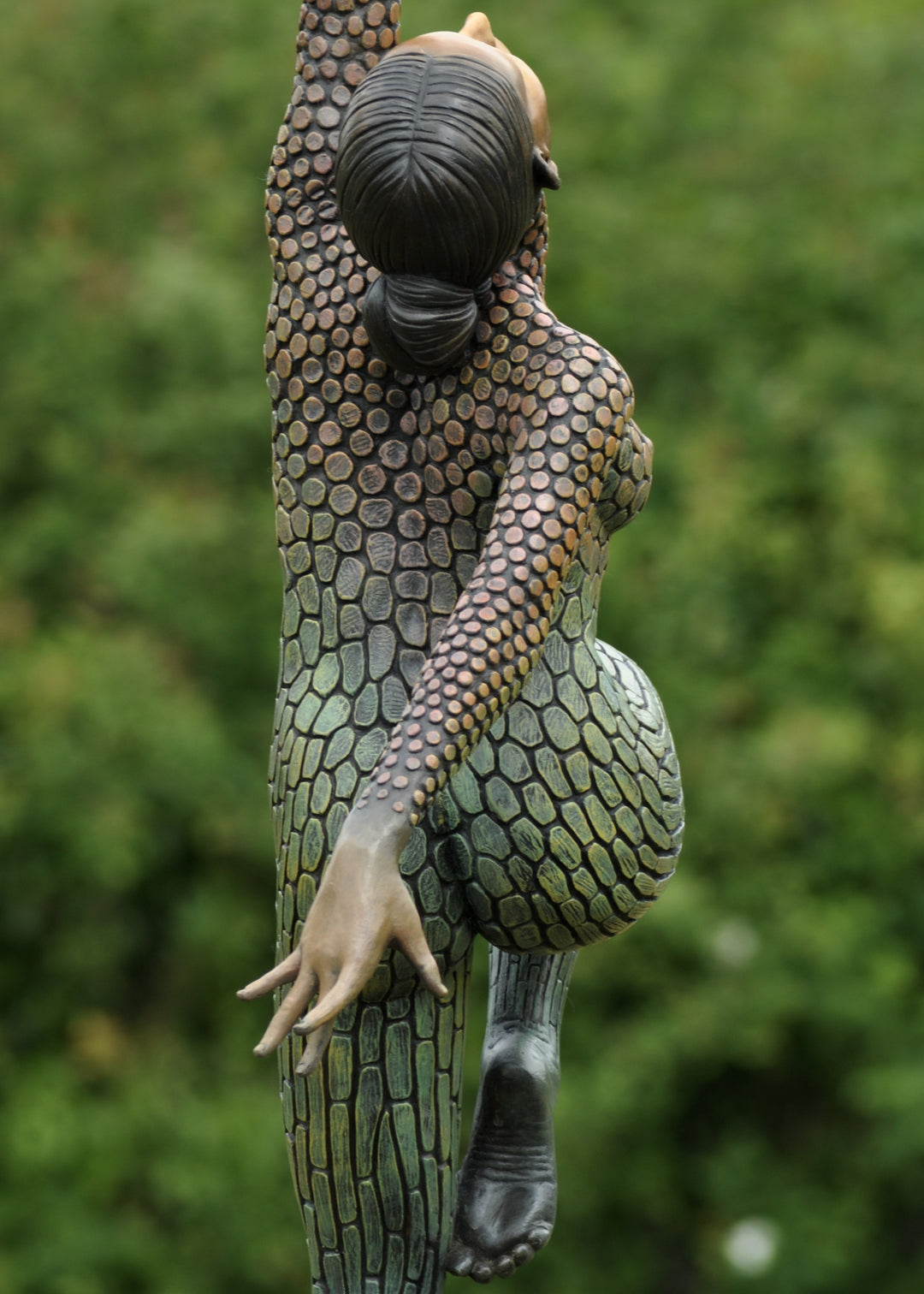 Life-size bronze sculpture of a woman balancing on one foot, her body covered in hand-painted mosaic-like textured scales that shift from earthy tones to green and blue patinas. She reaches upward with eyes closed, captured in a graceful, expressive pose against an outdoor garden backdrop.