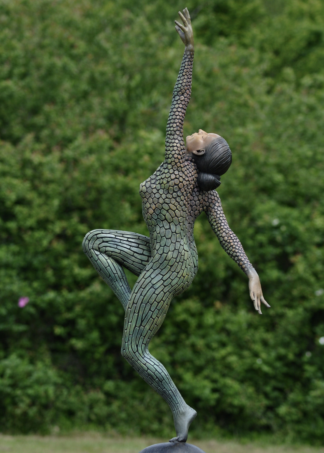Life-size bronze sculpture of a woman balancing on one foot, her body covered in hand-painted mosaic-like textured scales that shift from earthy tones to green and blue patinas. She reaches upward with eyes closed, captured in a graceful, expressive pose against an outdoor garden backdrop.