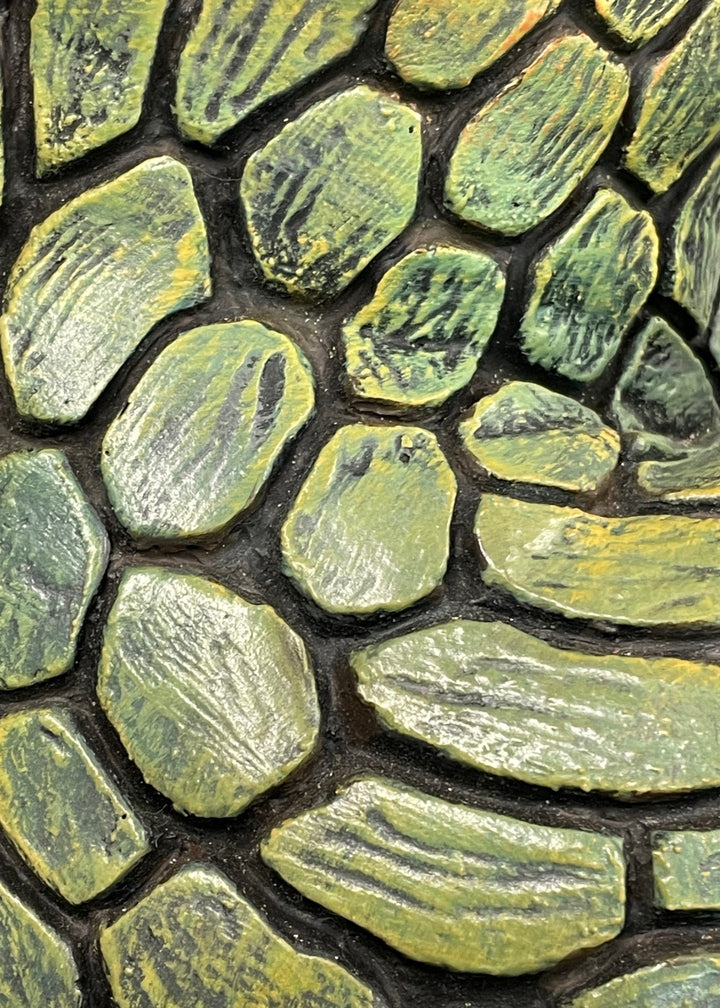 Life-size bronze sculpture of a woman balancing on one foot, her body covered in hand-painted mosaic-like textured scales that shift from earthy tones to green and blue patinas. She reaches upward with eyes closed, captured in a graceful, expressive pose against an outdoor garden backdrop.