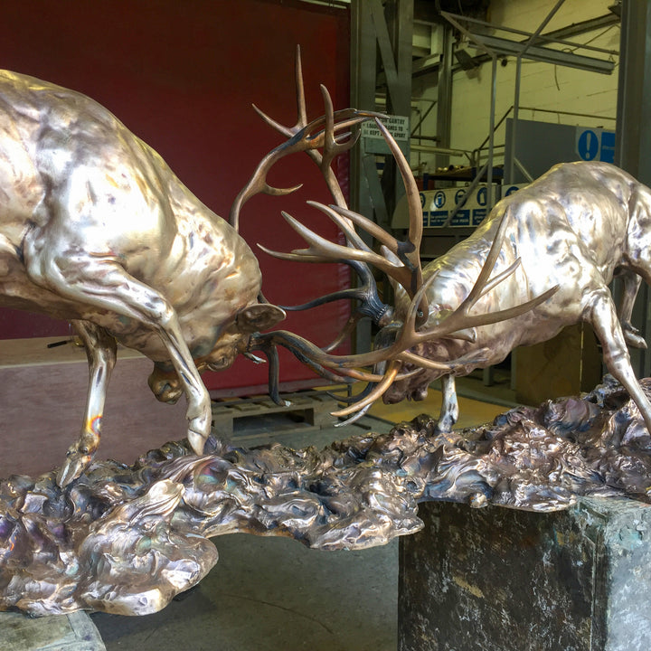 Bronze sculpture of two red deer stags in mid-rut, antlers locked in a dynamic head-to-head clash, highly detailed musculature and patina, mounted on a textured bronze base.