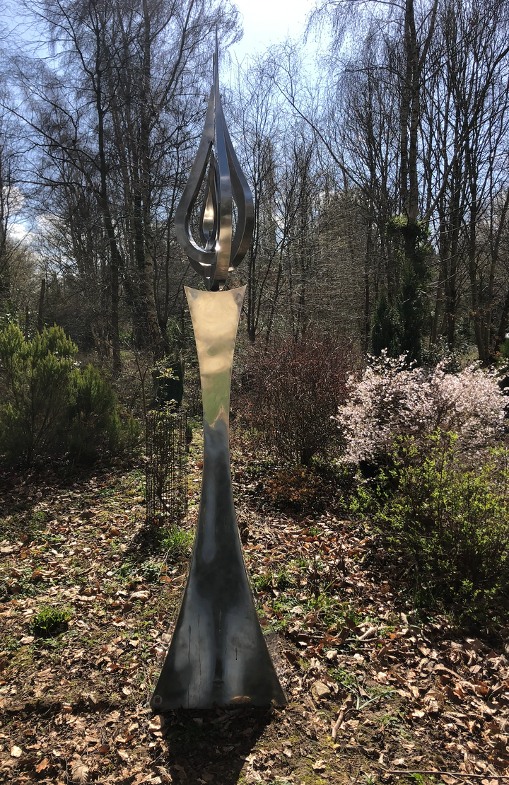 An elegant stainless steel kinetic sculpture by Martin Jarvis, designed in the form of a tall flame-like spire. The sculpture’s upper structure features fluid, ribbon-like curves that twist upward and move gently with the wind. Polished to a mirror finish, the steel reflects light and surroundings, giving it a dynamic presence. The base is solid and tapering, with soft surface patterning that adds depth and texture. Installed outdoors in natural and garden settings, the piece feels both modern 