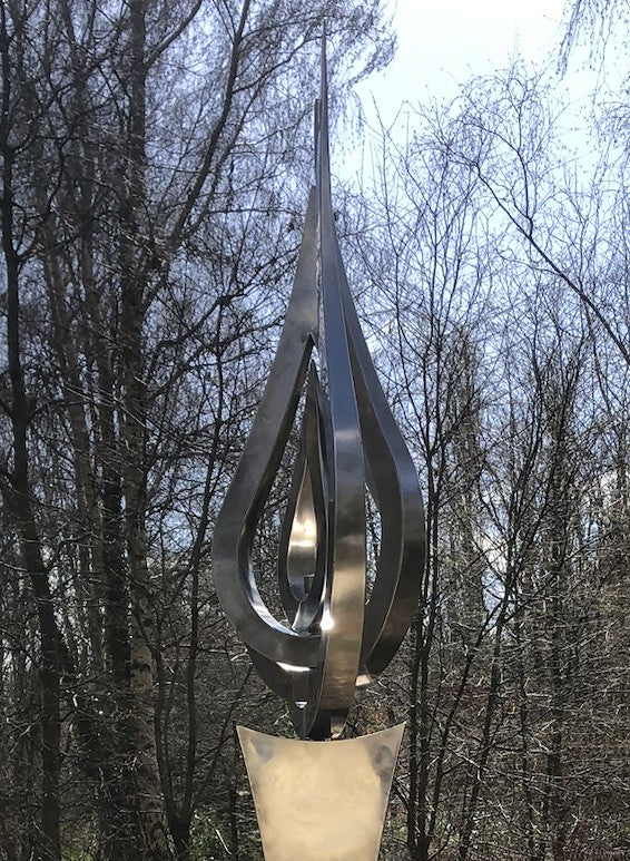An elegant stainless steel kinetic sculpture by Martin Jarvis, designed in the form of a tall flame-like spire. The sculpture’s upper structure features fluid, ribbon-like curves that twist upward and move gently with the wind. Polished to a mirror finish, the steel reflects light and surroundings, giving it a dynamic presence. The base is solid and tapering, with soft surface patterning that adds depth and texture. Installed outdoors in natural and garden settings, the piece feels both modern 