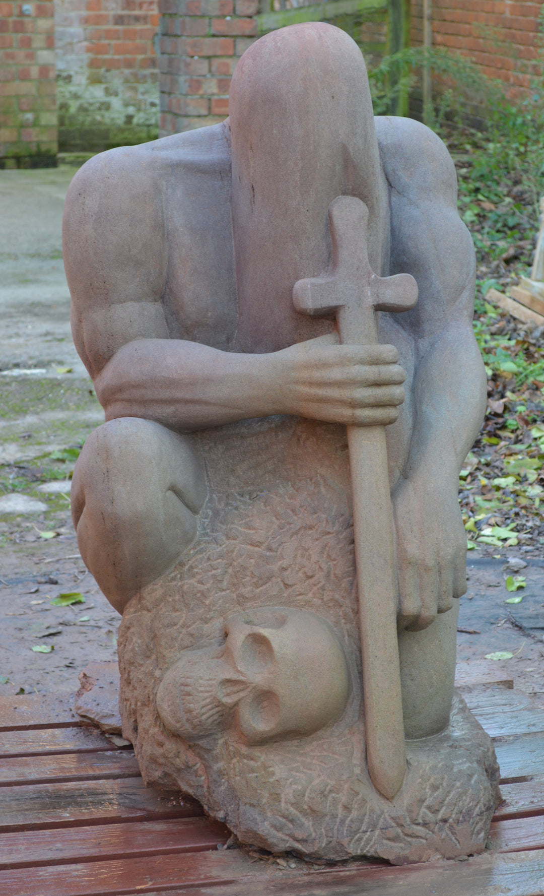 Deborah Harrison sculpture The Guardian, a monumental one-off sandstone figure kneeling with bowed head, gripping a sword planted into the ground. Carved from warm Forest of Dean sandstone, the faceless guardian form conveys strength, sacrifice, and timeless watchfulness, with a skull motif at its base adding symbolism of mortality and protection.